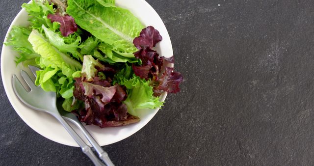 Image showing a white bowl filled with fresh green and red lettuce placed on a dark background. Perfect for use in health and wellness blogs, restaurant menus, diet-related articles, and promotions for vegan and organic food products.