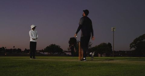 Dusk cricket bowler walking to stumps holding ball on community ground under floodlights