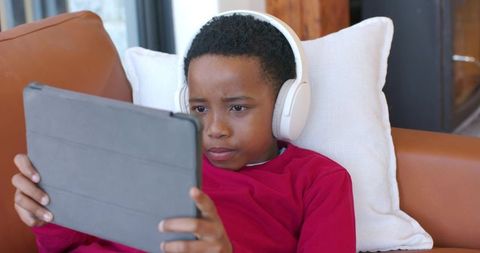 African american boy focusing on tablet while wearing white headphones and lounging on modern couch