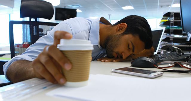 Man sleeping at desk wearing business casual clothes, holding takeaway coffee cup. Office environment includes computer, papers, and pens. Ideal for illustrating workplace stress, overwork, long hours, and fatigue in professional settings.