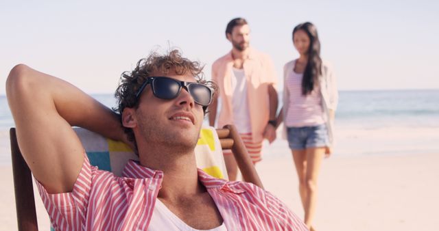 Young Man Relaxing on Beach Chair with Friends Walk in Background - Download Free Stock Images Pikwizard.com