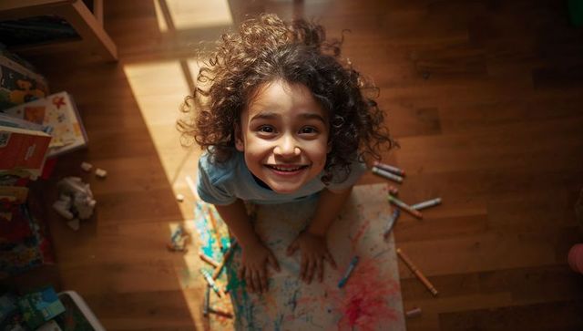 Sunlit curly-haired child drawing colorful crayon scribbles on wooden floor, smiling up