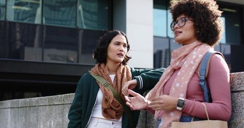 Young women chatting by city railing wearing scarves, backpacks and tote bags, casual urban lifestyl