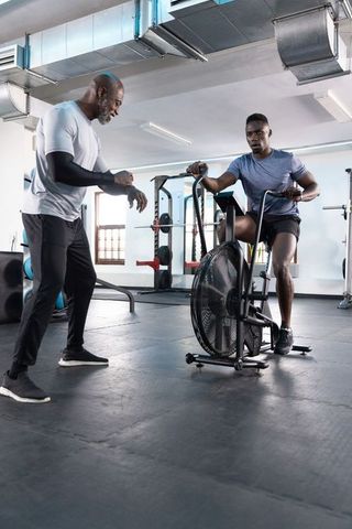 African american trainer coaching male athlete on fan bike near squat rack in modern gym