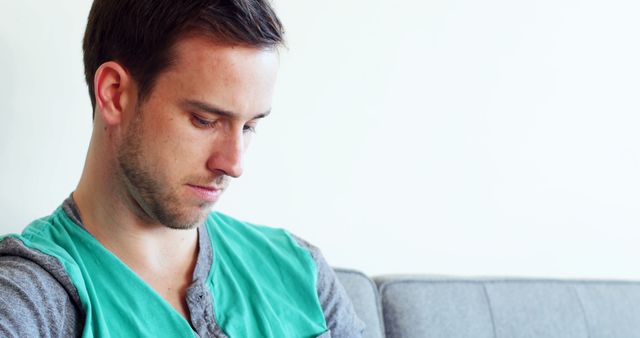 Young man sitting on a gray couch against light background, looking down with a thoughtful expression. Wearing casual, comfortable clothes. Perfect for articles about mental health, introspection, modern lifestyle, emotional wellbeing.