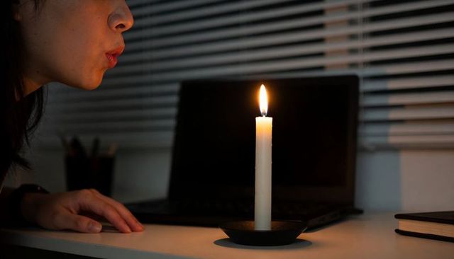 Woman blowing candle at desk with laptop moody nighttime warm candlelight glow