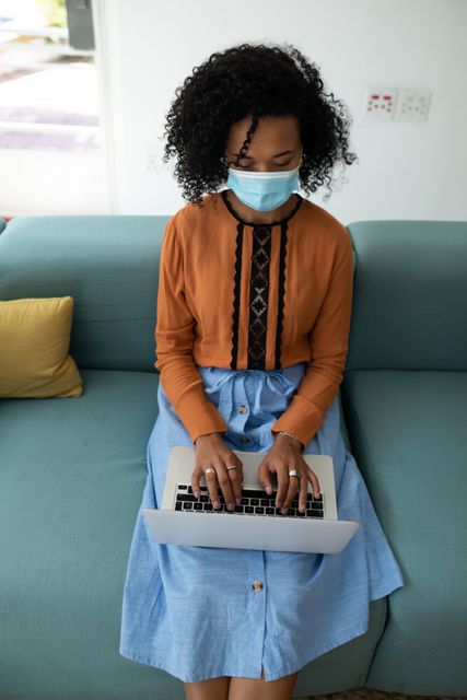African American woman wearing a facemask sitting on a couch in an office environment, typing on her laptop. She is dressed in casual attire, indicating a relaxed yet professional setting. This image can be used for topics related to remote work, pandemic safety measures, business productivity, and technology use in professional settings.