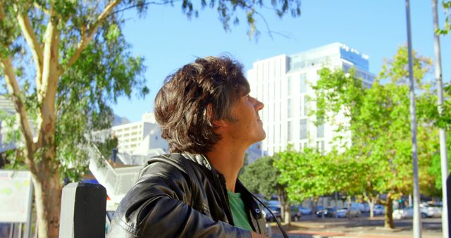 Young man with short hair wearing a leather jacket standing in an urban park area with modern buildings in the background and looking thoughtfully into the distance. Image useful for illustrating concepts of reflection, urban lifestyle, young adults, outdoor activities, and contemporary fashion.