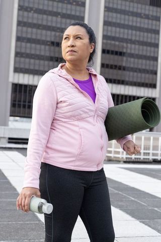 Determined mature woman holding yoga mat on urban rooftop in pink jacket and leggings
