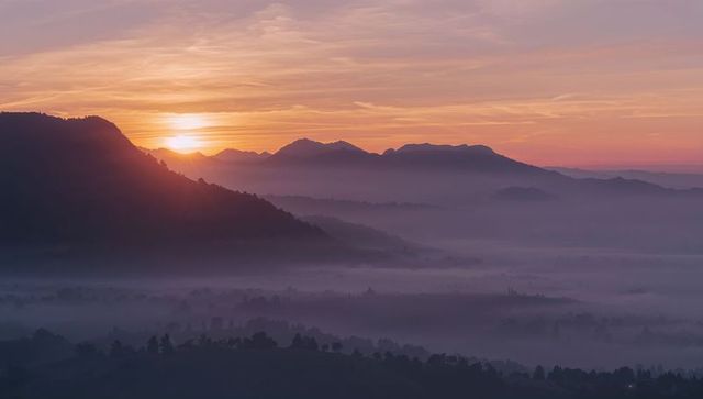Glowing sun peeking over misty alpine ridge, layering soft mountain valley at dawn
