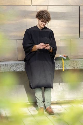 Graduate leaning on stone ledge, checking smartphone, cap and diploma nearby
