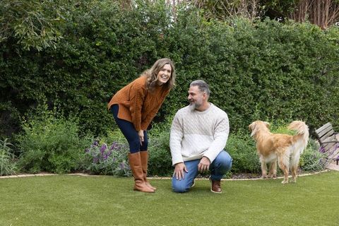 Couple kneeling on manicured lawn with golden dog, knit sweaters, backyard garden laughing