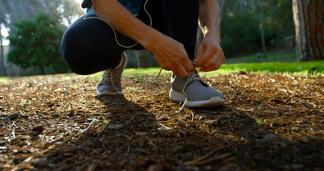 Unrecognizable person tying running shoes on a park's natural path. Suitable for topics related to fitness, outdoor activities, exercise, and healthy lifestyle. Ideal for illustrating preparation routines or promoting running and sports equipment.