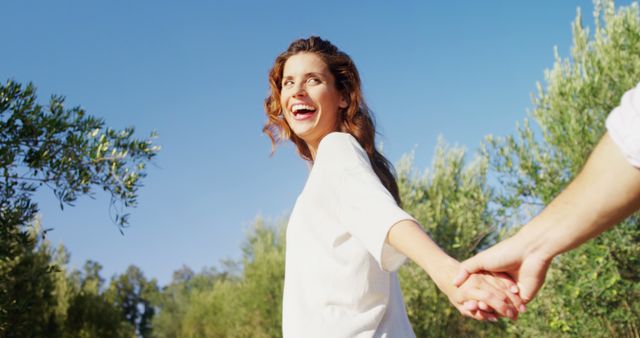 A young Caucasian woman is smiling joyfully while holding hands with someone outdoors, with copy space. Her expression and the sunny setting suggest a moment of happiness and connection.
