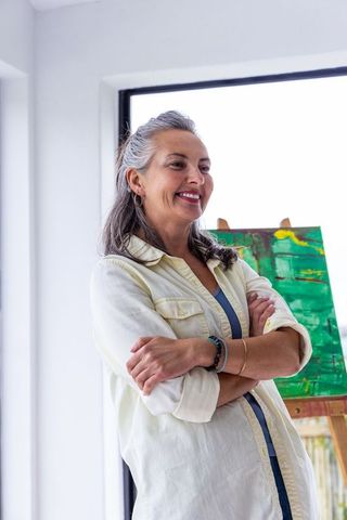 Mature female artist standing confidently in sunlit studio with abstract green painting