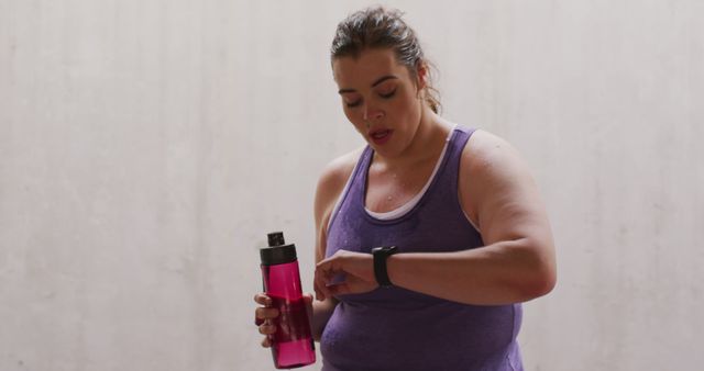 Woman wearing purple tank top holds water bottle while checking her fitness tracker. Suitable for themes related to fitness, technology in workouts, healthy living, and gym routines. Ideal for articles, blogs, advertising, and promotional material on exercise, sports, and health gadgets.