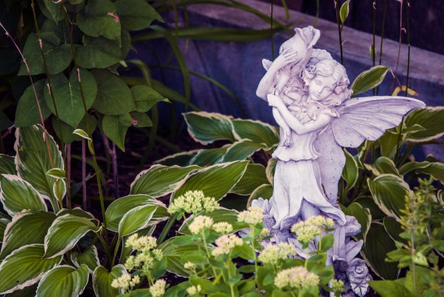 Fairy statue holding bird standing amidst lush green plants and foliage, evoking whimsical and serene garden feel. Ideal for use in content related to garden decoration, fairy tales, outdoor leisure, tranquility in nature, and landscape design inspiration.