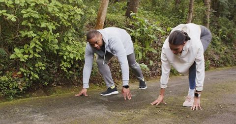 Diverse duo preparing for sprint on forest trail in hoodies and smartwatches