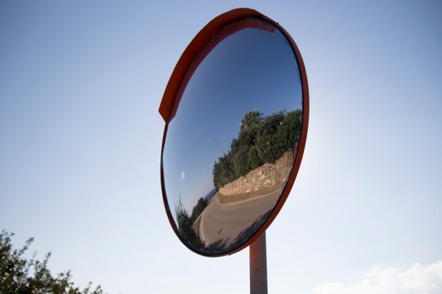Rural curved road reflected in a convex traffic mirror against a clear blue sky. Can be used for transportation safety articles, road sign visuals, or educational content about traffic mirrors.