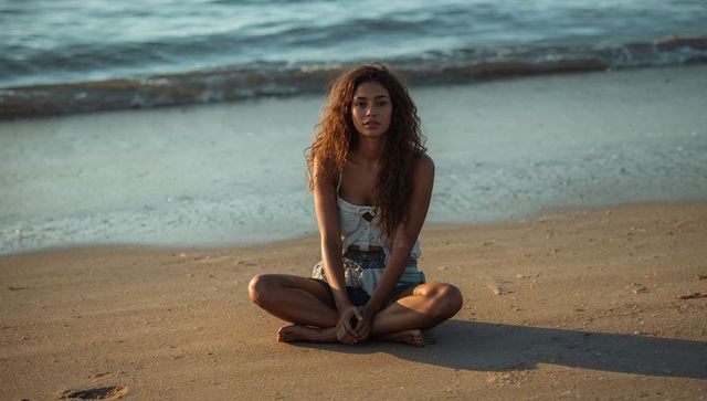 Woman sitting cross-legged on beach at golden hour wearing white crop top and denim shorts