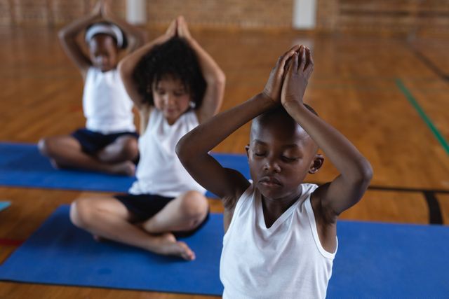 Schoolchildren sitting on yoga mats in a gym, practicing yoga and meditation. They are wearing white shirts and black shorts, with eyes closed and hands in prayer position above their heads. This image is ideal for use in educational materials, wellness programs, fitness campaigns, and articles promoting mindfulness and physical health in children.