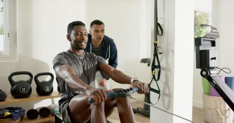 African american man rowing on ergometer while trainer coaching in bright home gym studio