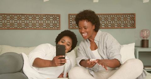 Two Women Laughing and Holding Smartphone While Relaxing on Cozy Green Living Room Couch