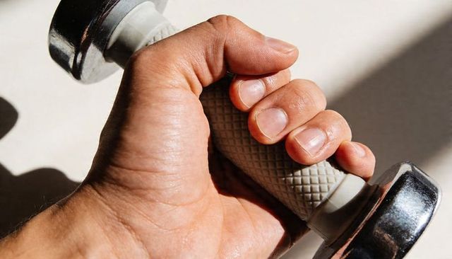 Gripping male-presenting hand lifting chrome dumbbell on pale surface with strong light