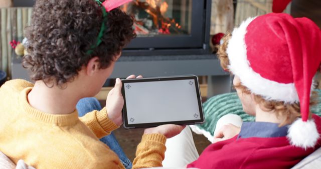 Two friends are celebrating Christmas, seated near a fireplace. One is wearing a Santa hat while holding a tablet. Perfect for festive advertisements, holiday greeting cards, and promoting digital devices during the holiday season.