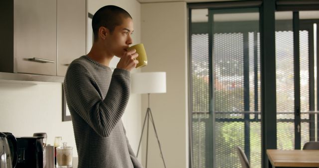 Young man sipping coffee in modern kitchen with large windows in the background, wearing casual grey sweater. Suitable for themes related to everyday life, morning routines, home interiors, modern lifestyles, and relaxation.