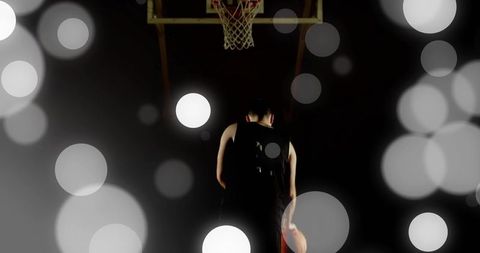 Rim-lit basketball player standing beneath hoop holding ball with moody bokeh silhouette