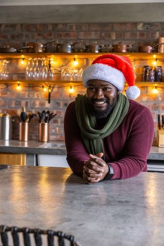 African american man leaning on kitchen counter wearing santa hat and scarf, smiling