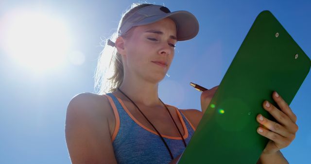 Shows a female coach assessing performance using a clipboard under bright sunlight. She is focused and appears focused and dedicated in a sporty outfit with a cap and whistle. Ideal for use in articles and advertisements about sports training, coaching, fitness programs, athletic events, or health and wellness promotions.