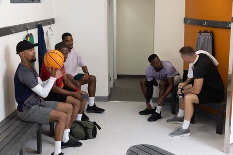 Multiracial basketball team sitting in locker room holding ball and talking before game