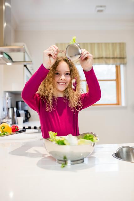 Young girl with curly hair preparing a fresh salad in a modern kitchen. She is smiling and holding a spoon with lettuce. Ideal for use in articles or advertisements related to healthy eating, family activities, cooking at home, and promoting a healthy lifestyle for children.