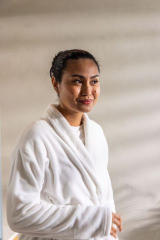 Asian woman standing in white plush bathrobe smiling and adjusting robe in sunlit interior