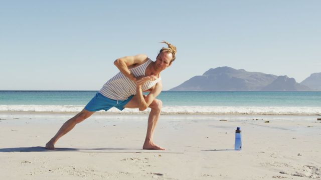 Man performing outdoor yoga on a sunlit beach, enjoying meditative practice with a dramatic mountain backdrop. Suitable for promoting fitness, wellness, beaches, healthy lifestyle content, travel destinations, and exercise gear.