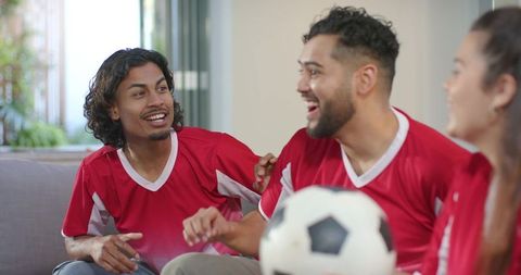 Diverse friends cheering while watching soccer match, holding ball on couch celebrating