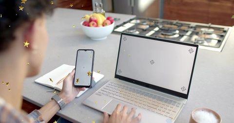 Man working from home holding smartphone and typing on laptop in kitchen with blank screens