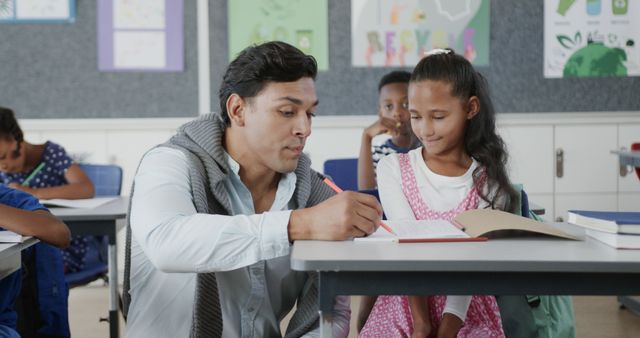Male teacher helping a young girl with her schoolwork in a diverse classroom. Background shows other children studying and colorful educational posters. Suitable for educational websites, academic blogs, diversity in education campaigns, and school promotional materials.