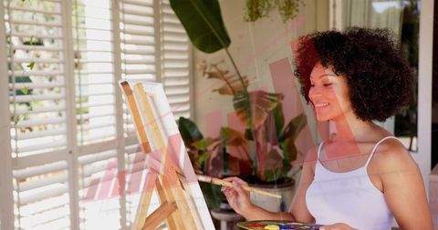 Sunlit artist painting on easel in bright home studio holding palette and brush smiling