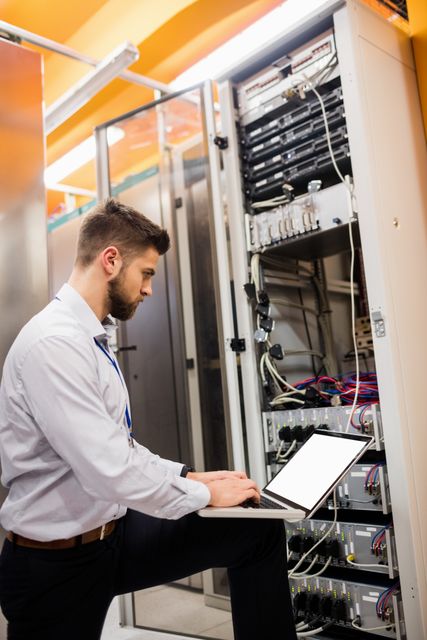This image shows a male technician using a laptop while analyzing a server rack in a data center. Ideal for illustrating concepts related to IT support, network management, server maintenance, and technological infrastructure. Useful for tech websites, IT service advertisements, and articles on computer networking.