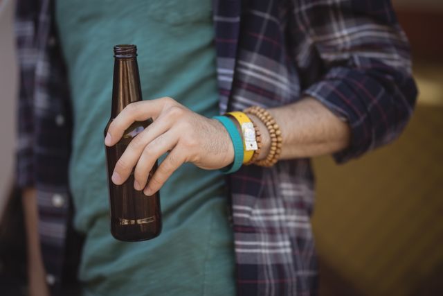Young man holding a beer bottle while standing outdoors. He is wearing a plaid shirt and multiple wristbands. This image can be used for themes related to casual lifestyle, leisure activities, outdoor gatherings, and youth culture.