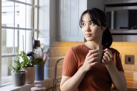 Asian woman standing by kitchen window holding ceramic mug and gazing at potted plants