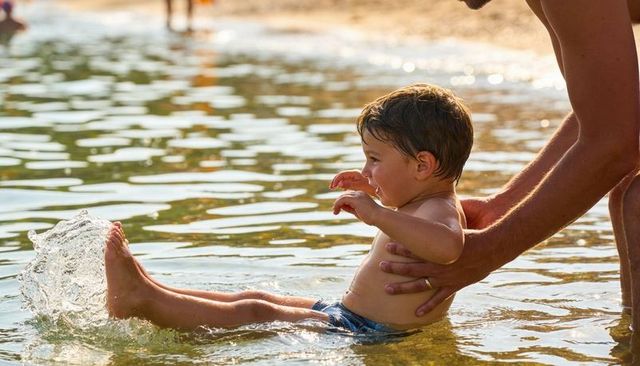 Smiling toddler splashing and kicking in shallow beach water with caring guardian hands