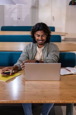 Young indian man smiling while working on laptop in modern coworking lounge teal wood interior