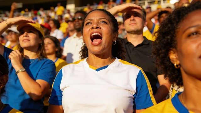 Stadium fan cheering in sunlight wearing blue yellow team jersey holding phone