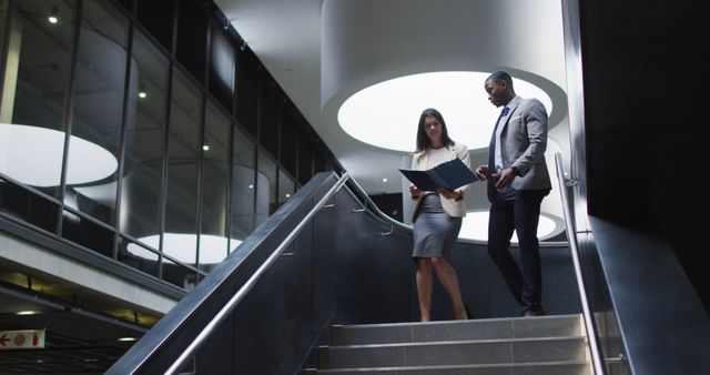 Depicts male and female business colleagues walking down a modern office staircase while discussing documents. Suggested uses include corporate presentations, business-related articles, teamwork promotions, and advertisements emphasizing corporate culture or professional collaboration.