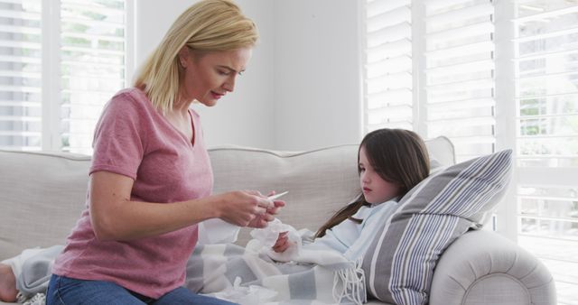 Mom is sitting beside her sick daughter on a couch, giving her medication. The setting appears to be a bright living room with white curtains, showcasing an attentive and caring atmosphere. Ideal for use in articles, healthcare promotions, parenting blogs, or family-oriented products and services.