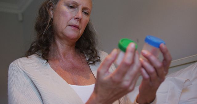 This image depicts a mature woman in a bedroom holding and inspecting small medical sample containers. The woman appears concerned or thoughtful. This can be used in articles or websites discussing personal healthcare, home medical testing, and the emotional impacts of medical concerns. Ideal for use in health blogs, patient care guides, or medical websites.